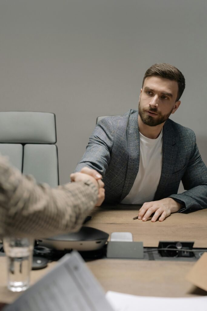 Two businessmen in a modern office shaking hands across a table. Professional setting.
